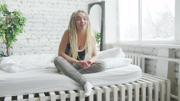 A Woman with Dreadlocks and Home Clothes Sits on a Large Bed and Talks to the Camera