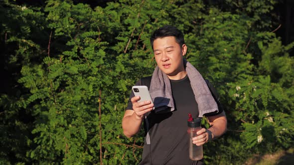Tired Satisfied Asian Sportsman Using Mobile Phone Drinking Water After Running