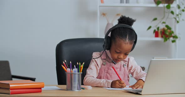 Concentrated Afro American Little Girl Child Sitting at Home at Table Draws Picture Chooses Pencil