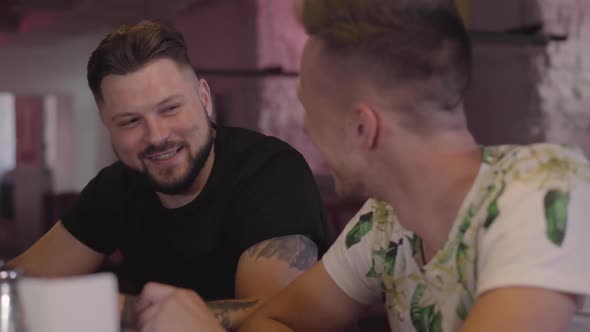 Two Adult Men Clink Glasses at the Bar Counter Smiling and Talking. Relaxed Male Friends Chilling