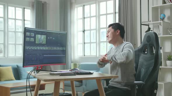 Asian Video Editor Man Stretching And Leaning Back While Using Computer For Editing Video At Home