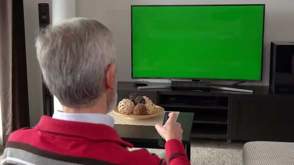 An Elderly Man Sits and Watches TV with a Green Screen and Switches Programs with a Remote Control