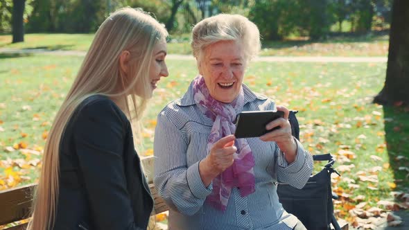 Beautiful Blonde Girl with Elder Woman Sitting on Bench and Looking Something on Smartphone