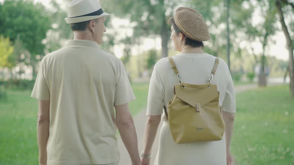 Back View of Relaxed Mid-adult Couple Strolling in Summer Park. Happy Caucasian Husband and Wife