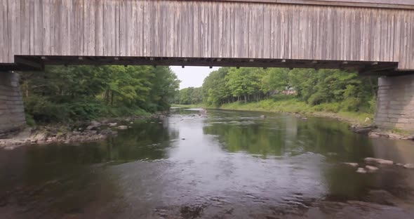 Aerial shot flying above a flowing river and underneath the covered wooden lowes bridge in the Maine