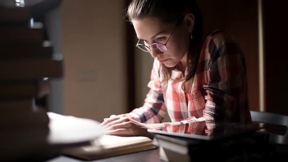 A girl reads a book while sitting late at a table in the light of a table lamp