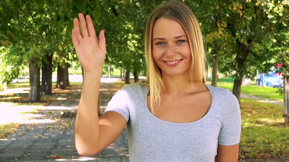 Young Pretty Blond Woman Waves with Hand - Park with Trees in Background