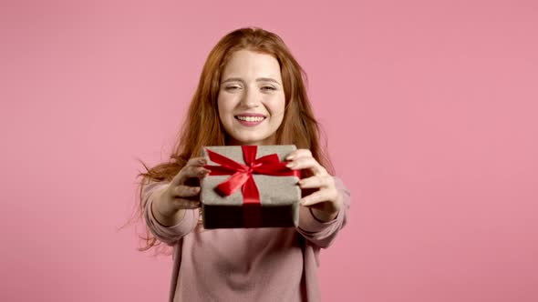 Cute Young Woman Holding Gift Box and Gives It By Hands To Camera on Pink Wall Background. Girl