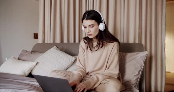 Young Beautiful Woman Using Laptop with Wireless Headphones Sitting in Cozy Bed