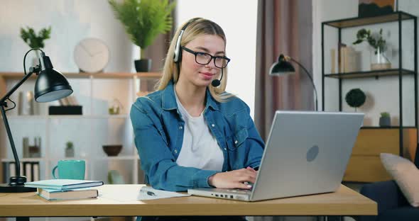 Young Blond Woman in Headphones which Sitting at Workplace at Home and Uses Computer