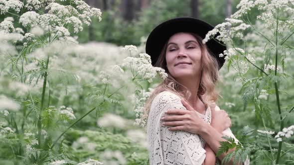 Young Woman in a Hat Among White Flowers in the Forest, Enjoying Nature and Freshness in Summer