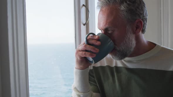 Caucasian mature man looking through the window and drinking coffee