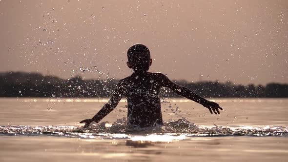 Silhouette of Happy Boy at Sunset Creating Splashes of Water with His Hands. Slow Motion