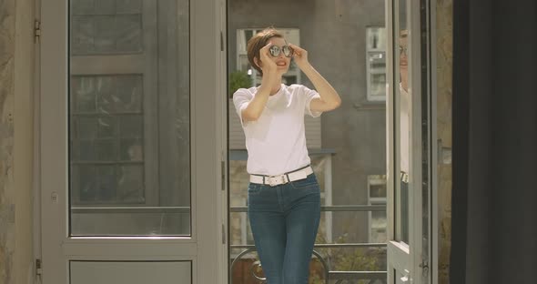 Portrait of Joyful Young Caucasian Woman Standing on Balcony and Putting on Sunglasses. Positive