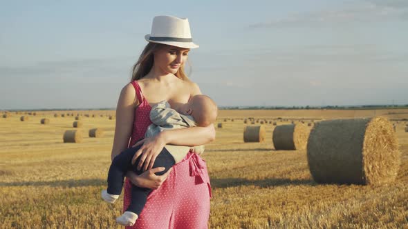 Happy Young Mother in Hat Feeds Baby Son with Breast in Haystack Field in Summer