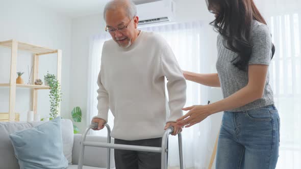 Asian young daughter support senior older man walk with walker at home.