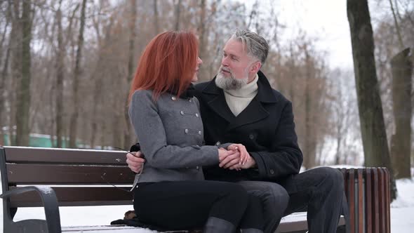 an adult gray-haired man sits on a bench in the park with his girlfriend