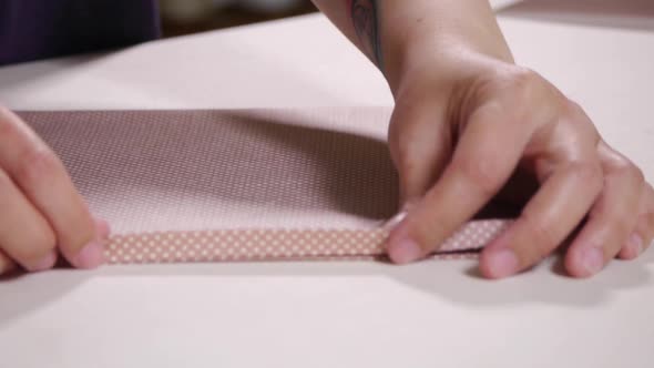 Fingers folding and unfolding edges of light pink colored fabric on white table