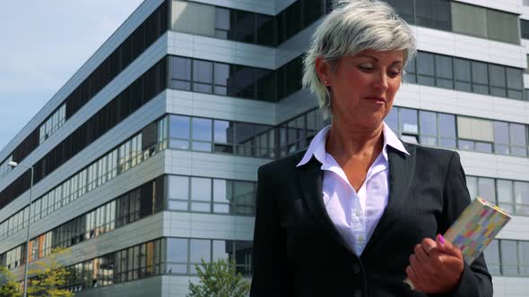 Business Middle Age Woman Holds Book in Hands and Smiles To Camera - Company Building in Background