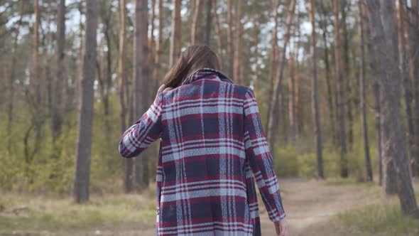 Camera Following Young Caucasian Woman in Plaid Jacket Walking in Forest. Cheerful Cute Girl