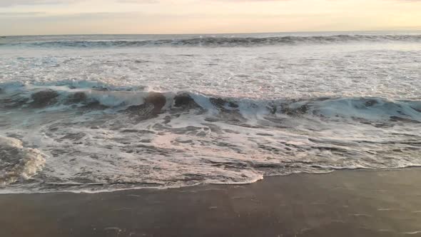 Aerial forward over waves crashing at sandy beach in front of sunset horizon line