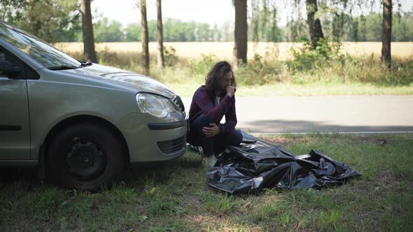 Young Male Driver Crying Holding Head in Hands Sitting at Car and Corpse Covered with Sheet Outdoors