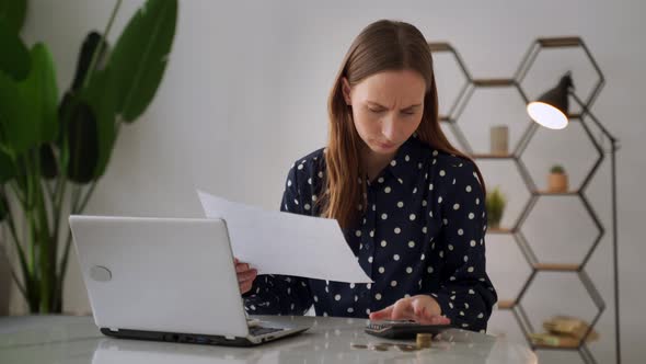 Worried Woman is Sitting at a Desk Using a Laptop Checking Finances Using a Calculator