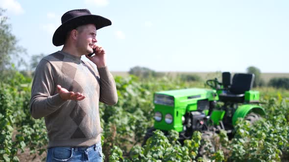 A Young Rural Farmer is Talking on the Phone on a Vineyard with a Tractor Near Him