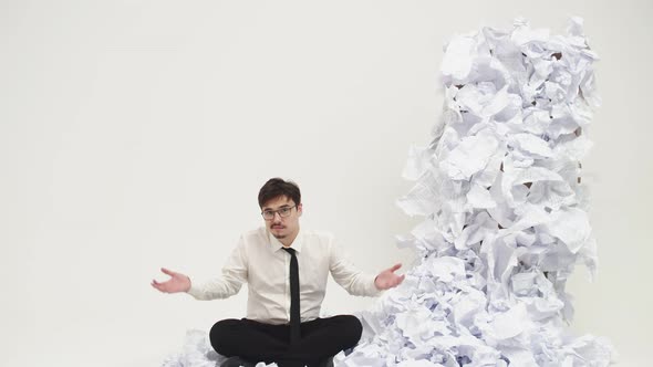 Male office worker sits on floor and spreads hands not knowing what to do next to mountain document