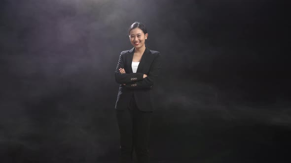 Smiling Asian Speaker Woman In Business Suit Crossing Her Arms While Standing In The Black Studio