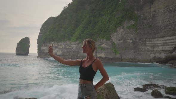 Charming blond woman taking selfies at tropical beach with tall cliffs in background, joyful vacatio