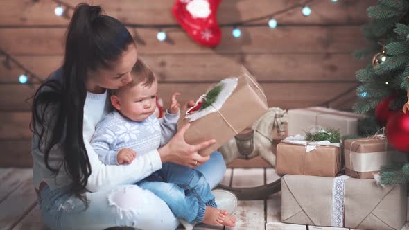 Young Brunette Mother and Her Little Baby Wrapping Gifts Under Christmas Tree at Home at Xmas Eve