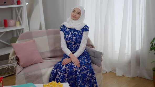 Charming Young Slim Middle Eastern Woman in White Hijab and Blue Wedding Dress Sitting on Couch