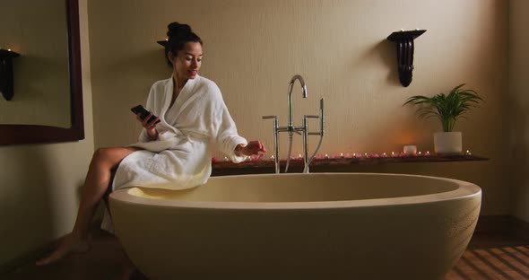 Happy biracial woman sitting in robe on bathtube, preparing bath, using smartphone