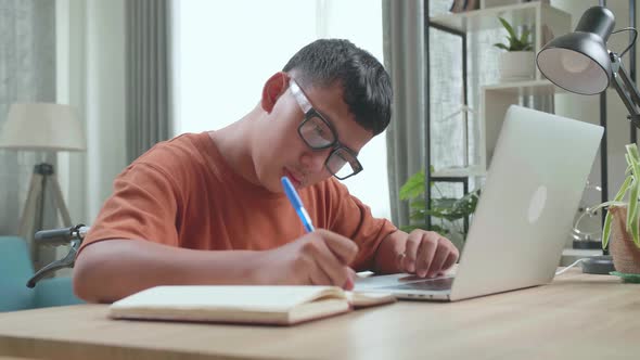 Young Asian Boy Sitting In A Wheelchair While Using Laptop Computer And Note In Book At Home