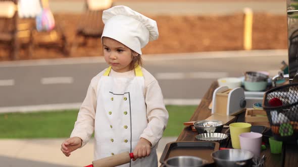 Adorable Little Girl in Chef's Coat and Cap Cooks at the Children's Toy Kitchen