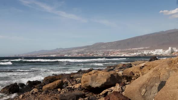 View of the Atlantic waves coming to the rocky shore with a city in the background, Tenerife