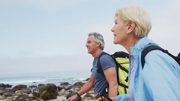 Senior hiker couple with backpack and hiking poles walking while hiking on the beach.