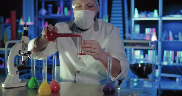 Scientist Chemist in Protective Glasses Pours the Solution Into a Glass Beaker Shakes It and Looks