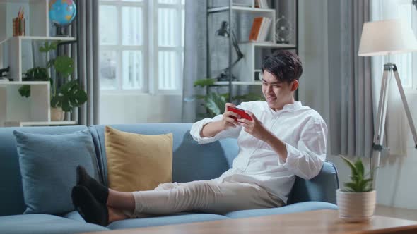 Asian Man Playing Game On Smartphone And Smiling While Lying On Sofa In The Living Room