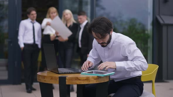 Portrait of Busy Bearded Man with Laptop and Tablet on Office Terrace with Blurred Colleagues