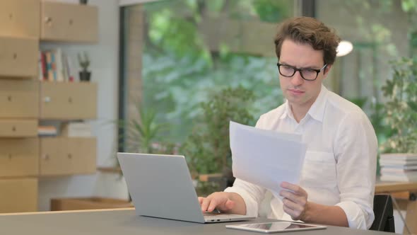 Man with Laptop Reading Documents in Modern Office