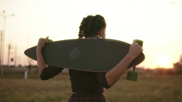 Back View of Woman in Plaid Shirt and Tank Top Holding Longboard and Walking During the Sunset in