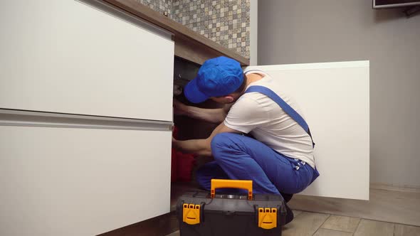 plumber in overalls and white T-shirt examines pipes in kitchen in apartment.