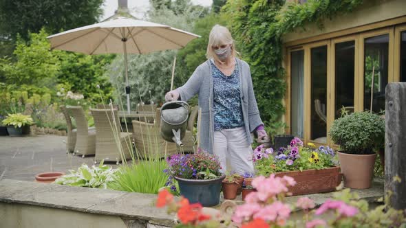 Elderly woman watering flower pot in backyard wearing face mask during coronavirus quarantine