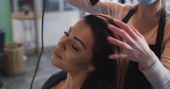 Female hairdresser wearing face mask blow drying hair of female customer at hair salon