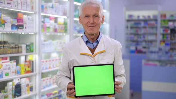 Medium Shot of Smiling Senior Male Pharmacist Showing Digital Tablet with Green Screen Looking at