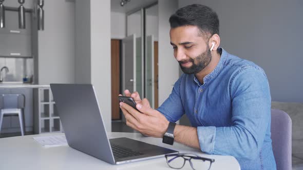 Young Adult Smiling Indian Businessman Holding Phone Sitting Front of Laptop