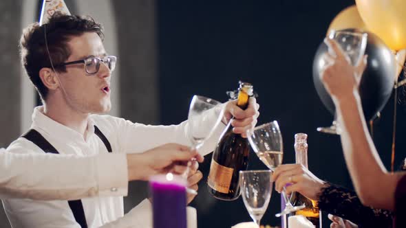 Millennial Friends at Party Sitting at Table Opening Bottle of Champagne and Pouring Sparkling Wine