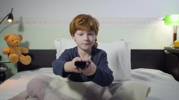 Front View Portrait of Cute Caucasian Redhead Boy Sitting on Bed and Switching Channels with Remote